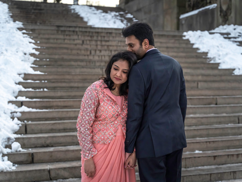 A couple stands in front of the steps of the Bethesda Terrace with snow in the background.