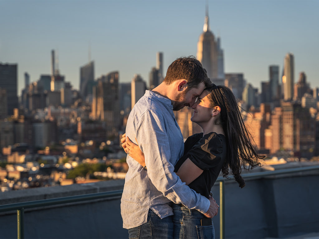 Jordan Zauderer and his fiancé, embrace each other during golden hour after their West Village rooftop engagement with the Empire State Building in the background.