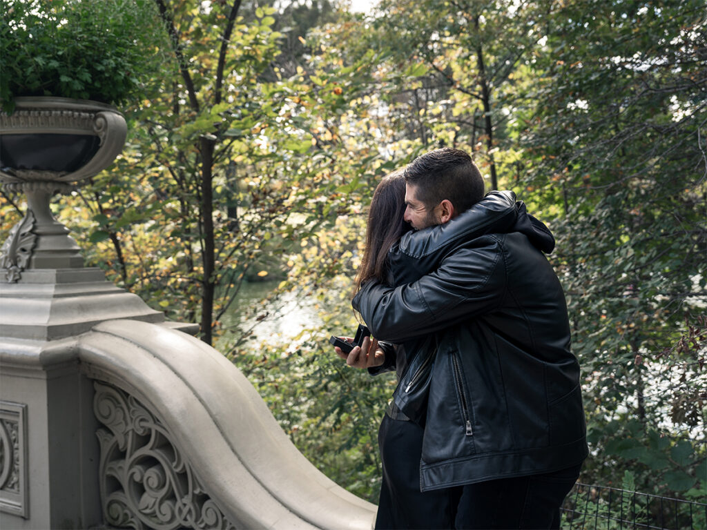 After popping the question during a surprise proposal at Bow Bridge in Central Park, the engaged couple embrace in happiness.