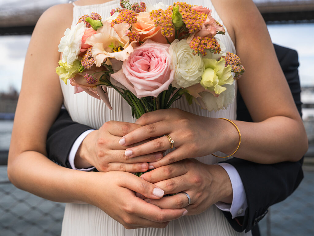 Detail shot of a couple holding hands and the brides bouquet, while also showing their newly wedded status with their wedding rings.