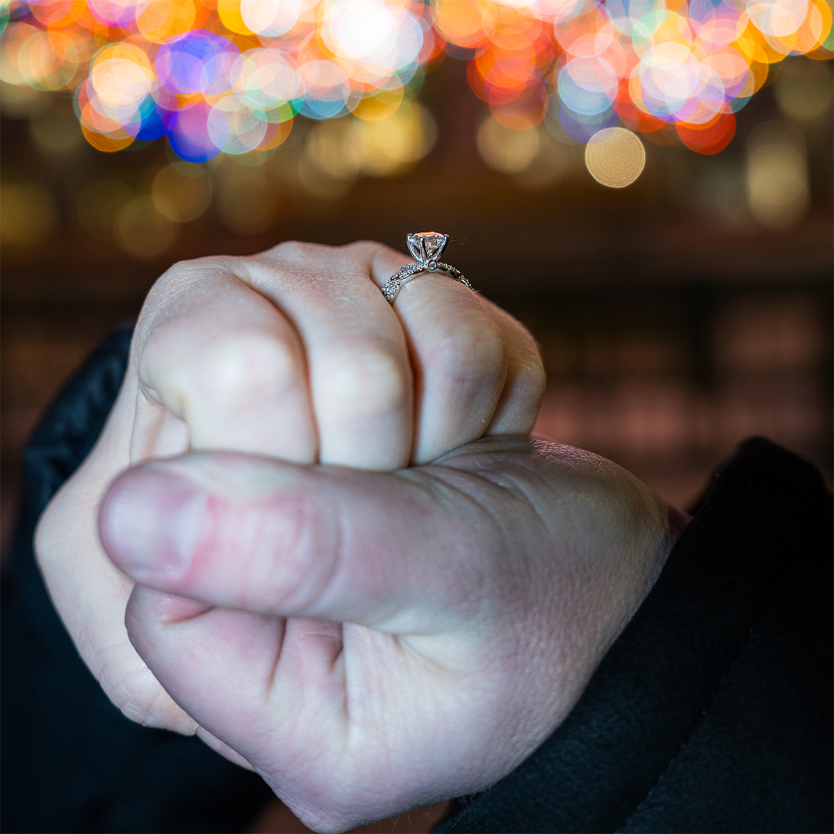 Newly engaged couple clasps hands, showing off the diamond ring against the bokeh of the colorful Rockefeller Center Christmas Tree lights in the background.