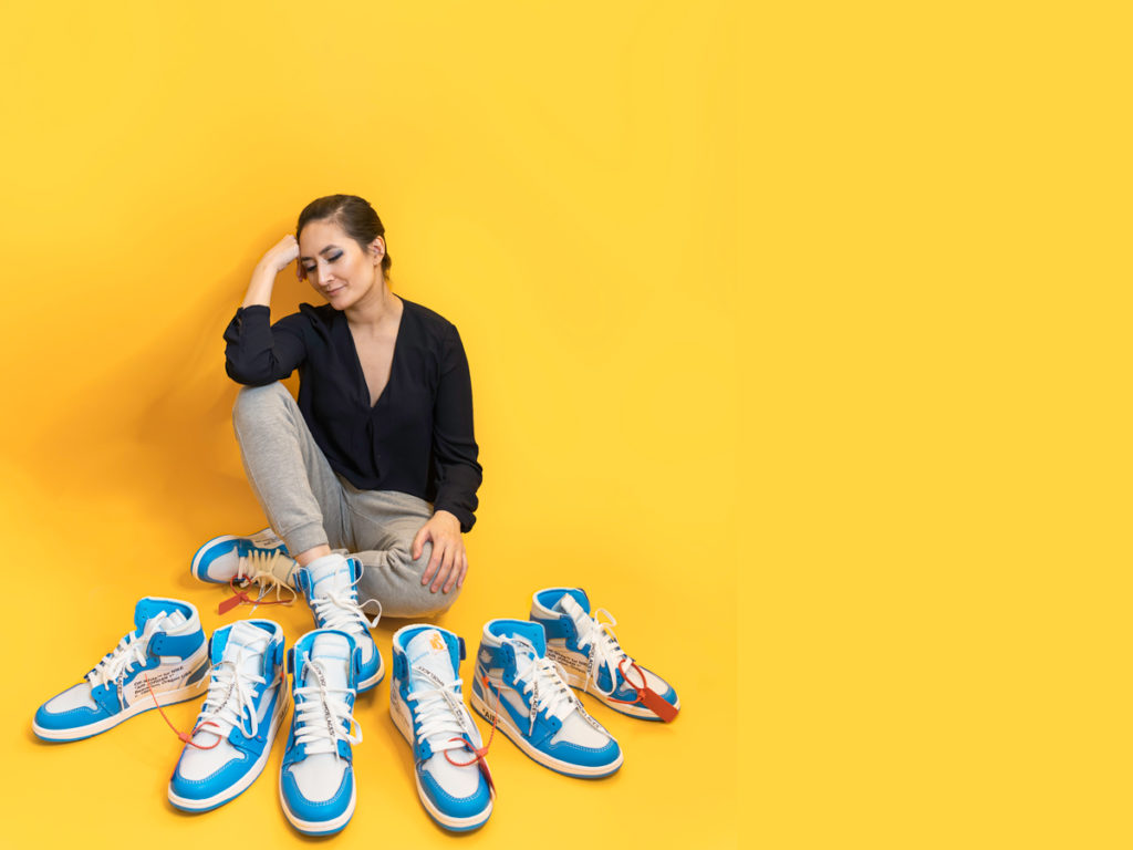 Self portrait content creation of Erin Donahue sitting on the ground with four pairs of Nike Off-White sneakers and a yellow background.