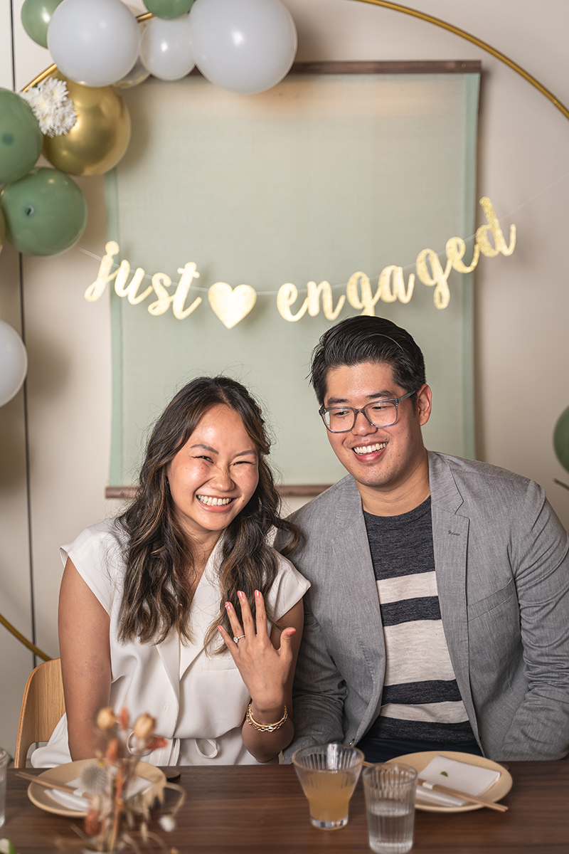 Tom and Janet sitting in front of the "just engaged" sign in a private room at Rule of Thirds, Brooklyn and showing of their engagement ring.