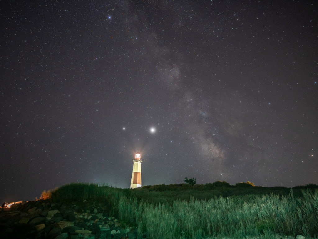 Light painting photography of the foreground at Montauk Lighthouse illuminates with the Milky Way in the background.