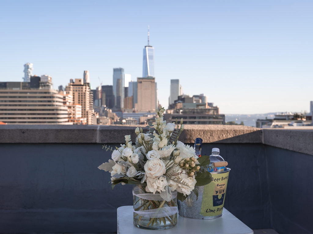 A bouquet of flowers and a bucket of drinks sits on a table with the view of the World Trade Center in the background for the perfect West Village rooftop engagement.