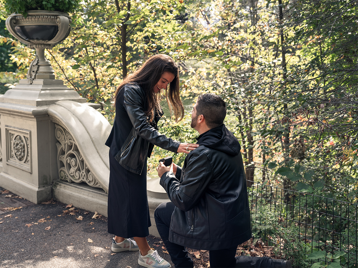 Groom-to-be gets down on one knee in front of the Bow Bridge in Central Park to propose to his girlfriend.